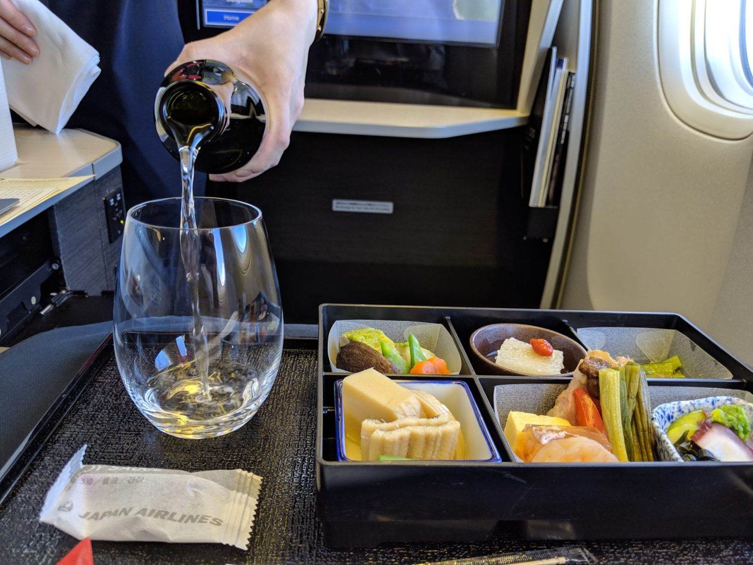 a flight attendant pours sake into a wine glass next to a tray full of food