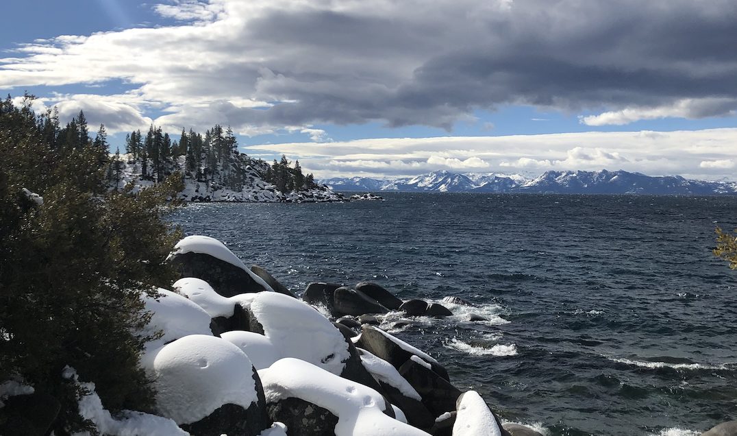 The snowy, rocky banks of Lake Tahoe with the Sierra Nevada mountain range on the horizon. 
