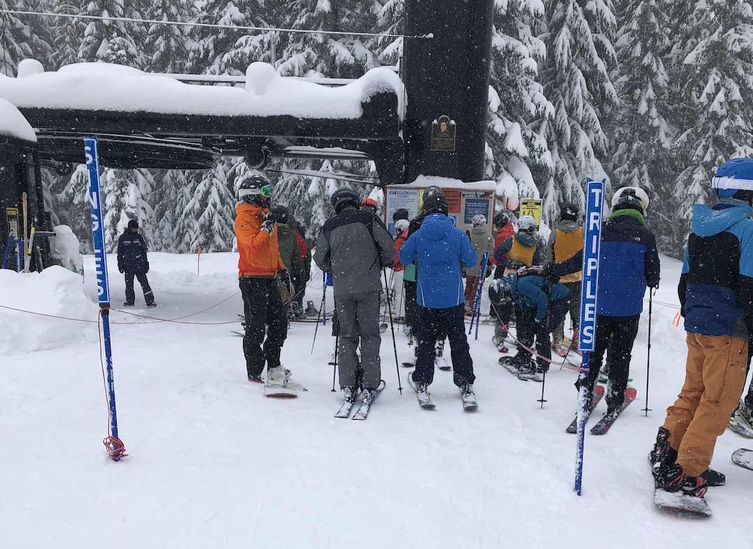 Skiers line up at an old chairlift while the snow falls on a powder day. The lines delineate where "singles" and "triples" should line-up. 