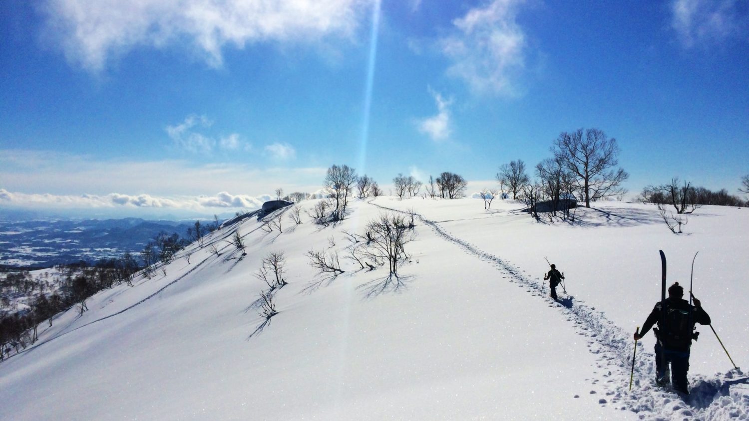Two skiers hiking through deep snow on top of a mountain in Niseko, Japan on a bluebird day. 