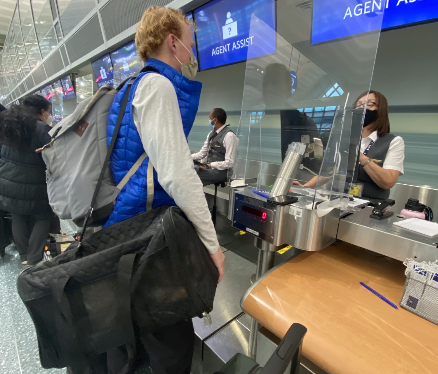 A traveler in a blue vest checks in with a Delta agent carrying a black pet carrier with a small dog inside. 
