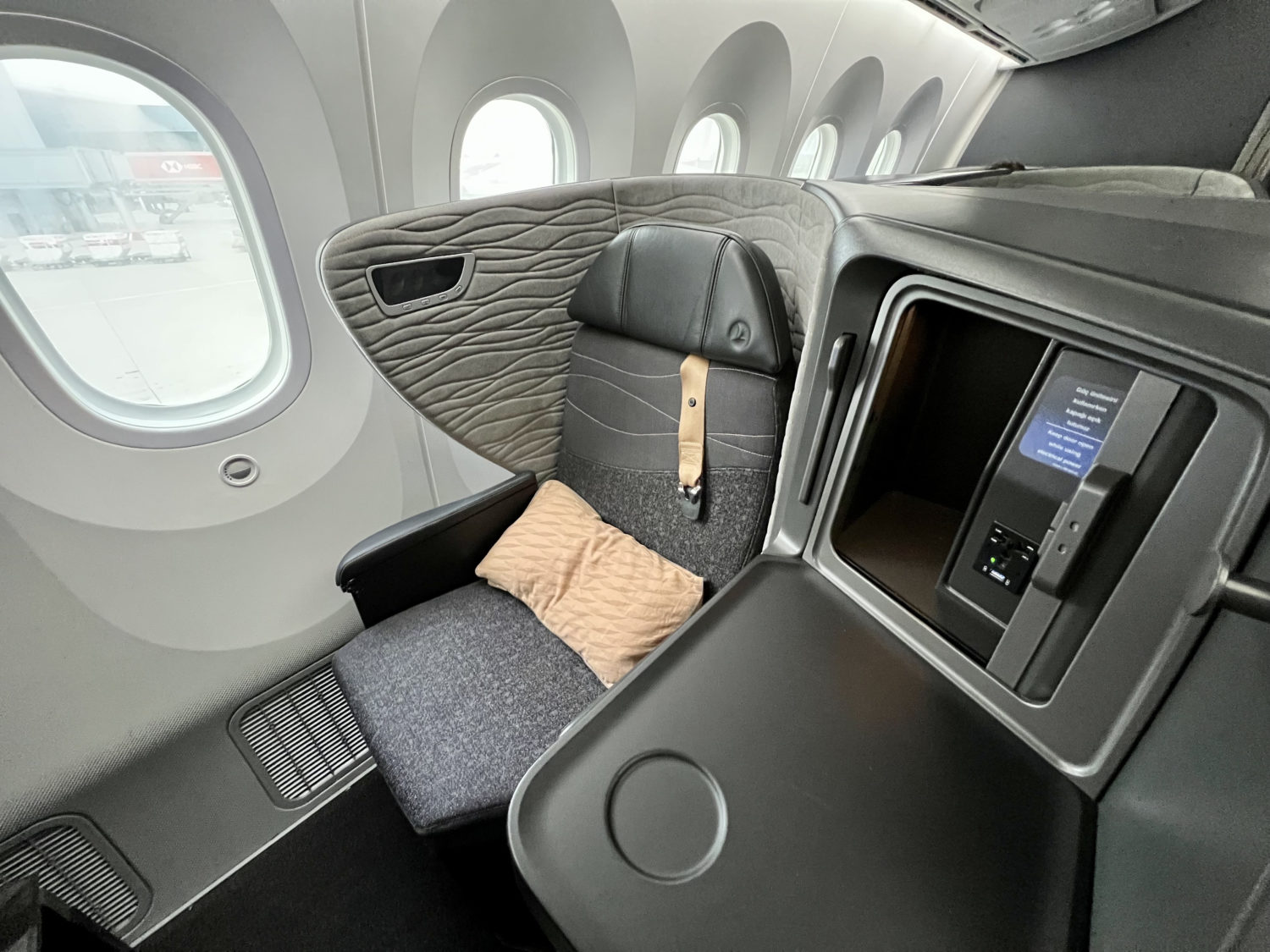 a solo business class seat with gray cushions and gray walls and a console table. A brown pillow is on the seat. 