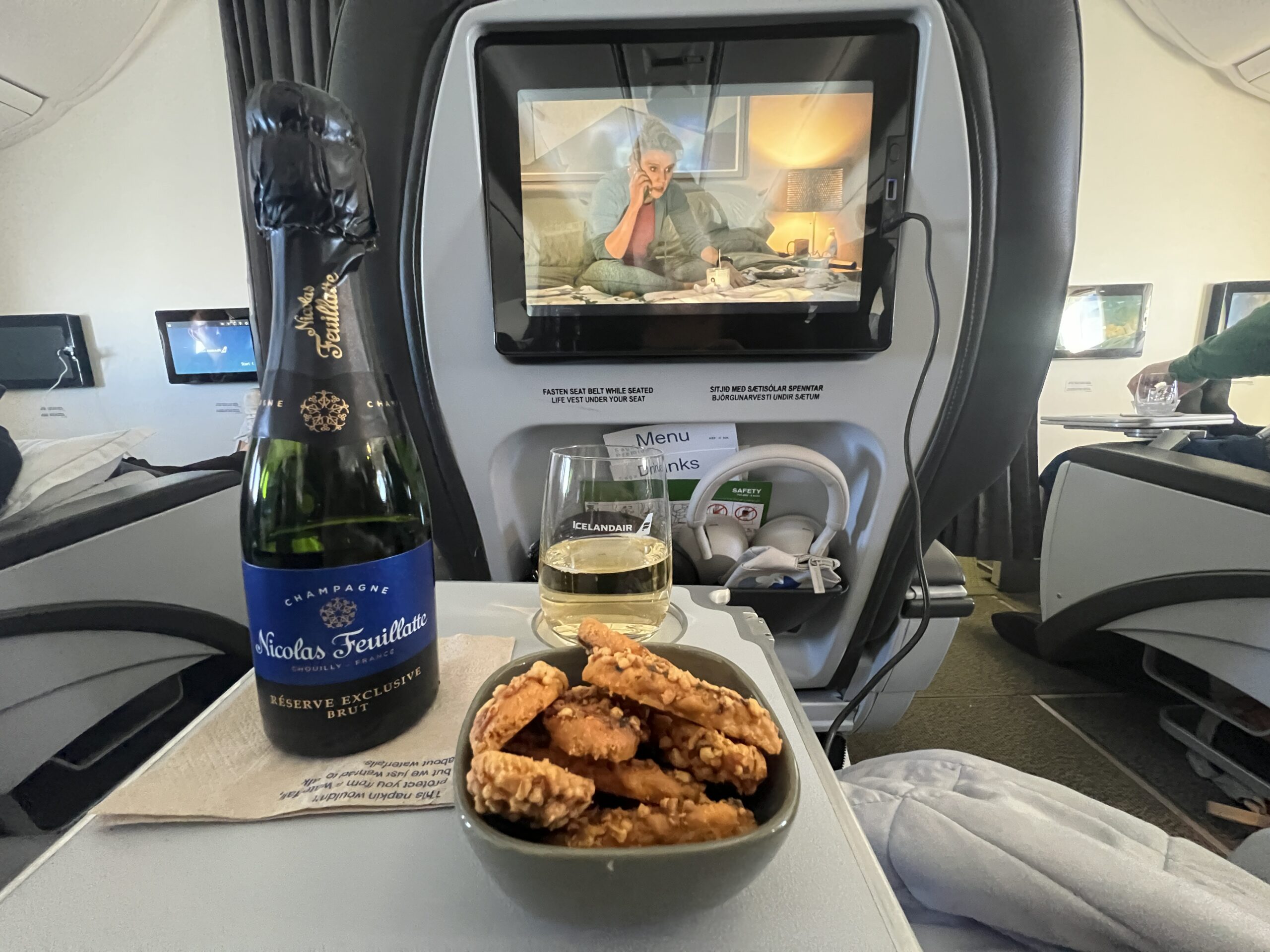 An airplane tray table holding a mini bottle of champagne, a glass of champagne, and a bowl of cheese crisps. A movie is playing on the seat back screen in the background. 