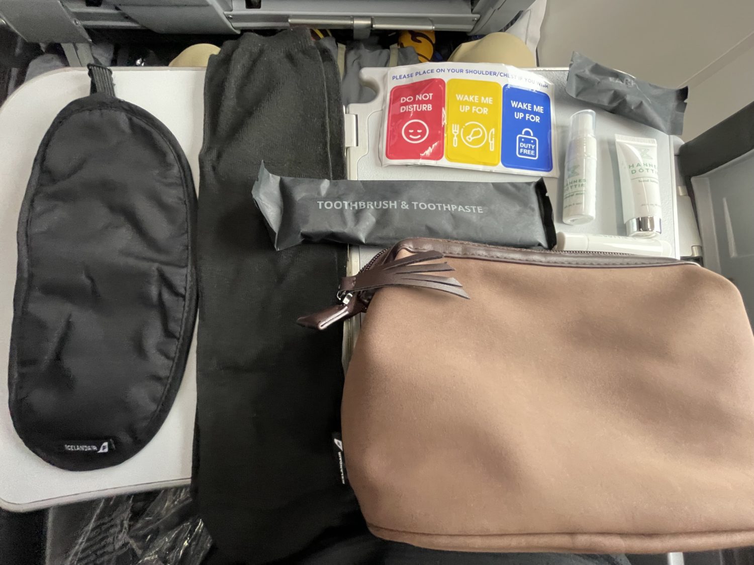 The contents of an amenity kit sprawled out on a tray table. An black eye mask, a black pair of socks, toothbrush and toothpaste, lotion, and a brown pouch