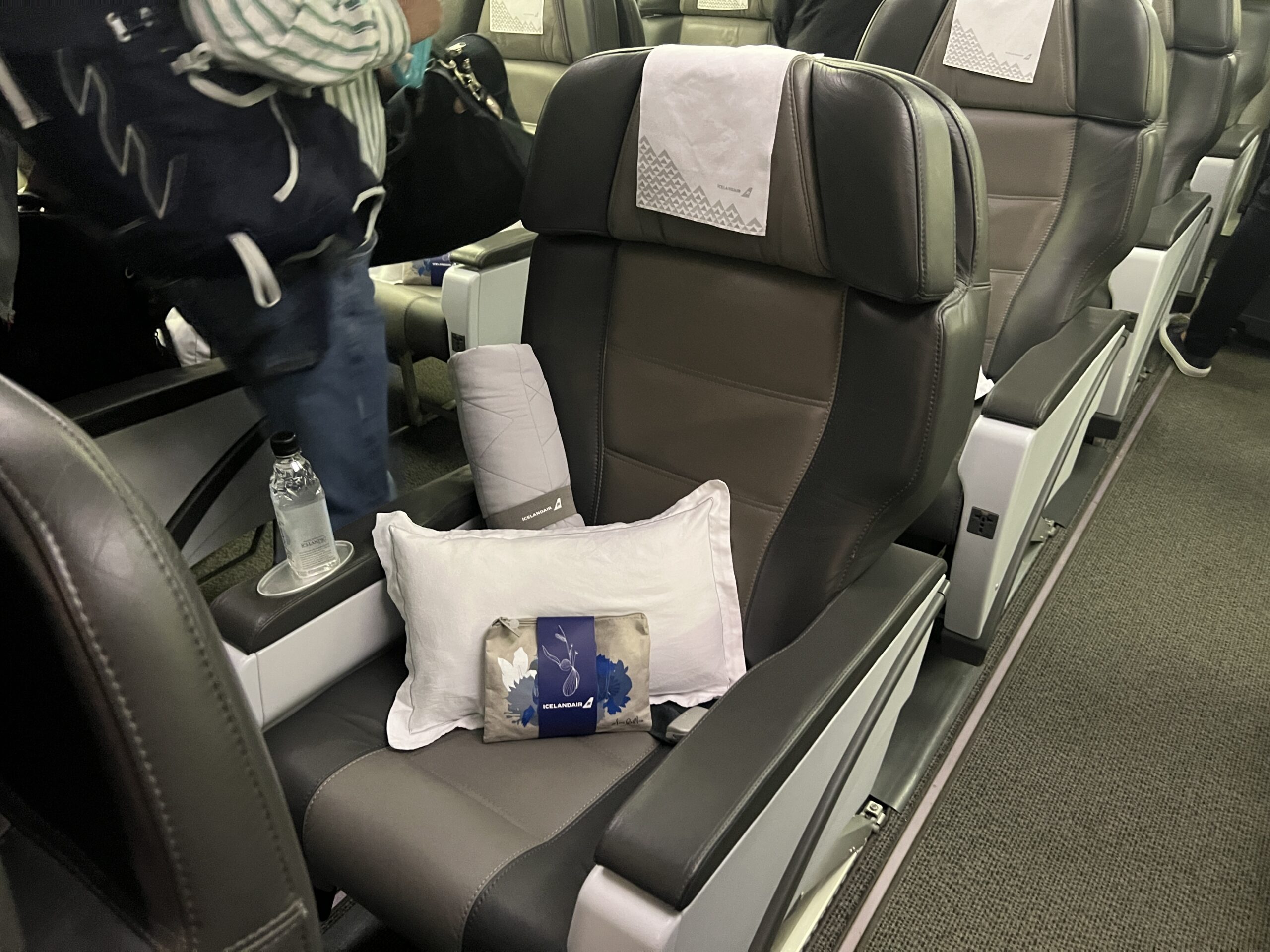 Middle seat in business class. At the seat is a water bottle, pillow, blanket, and floral pouch with amenities