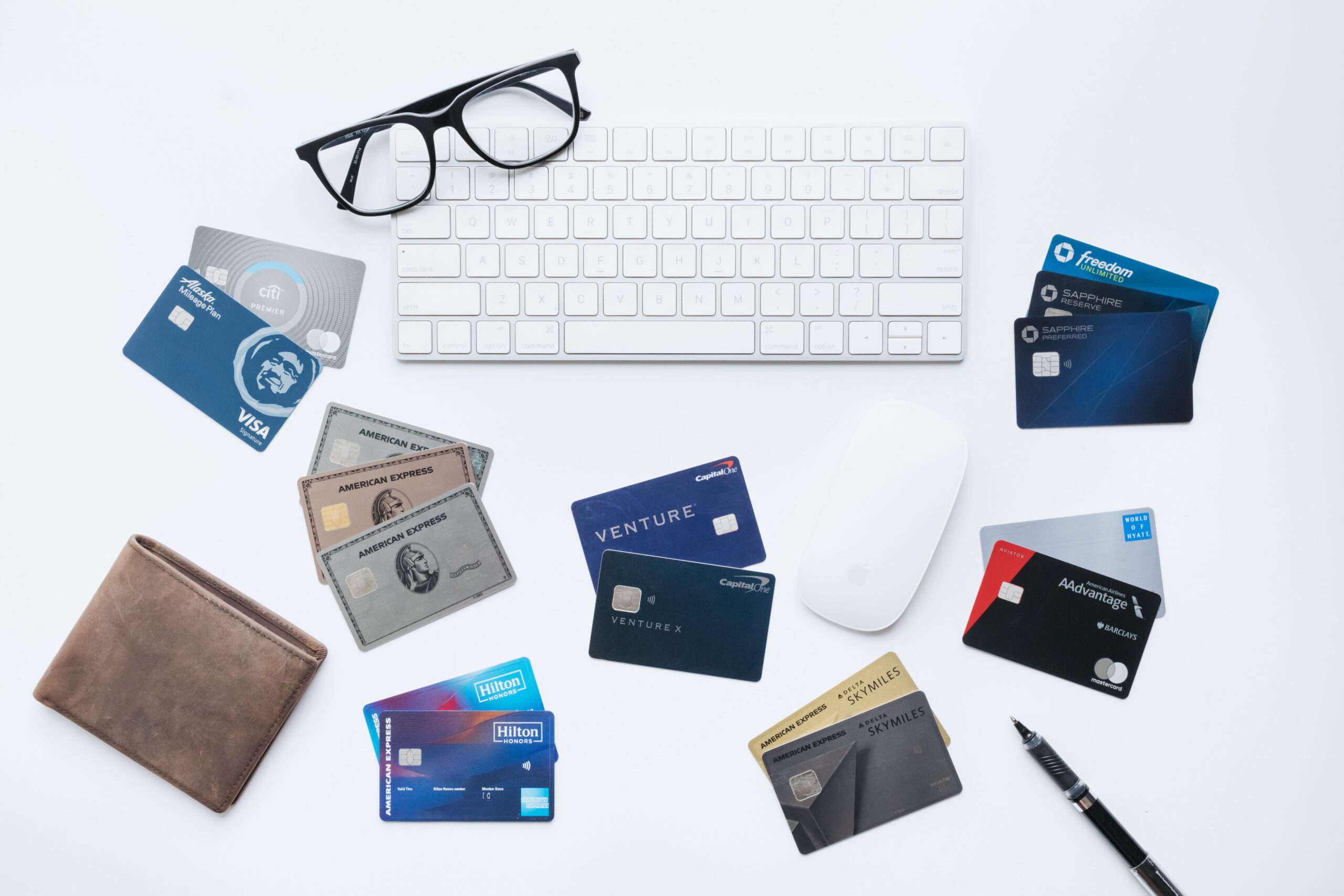 Credit cards spread out on a desk with a pair of glasses, a wallet, keyboard, and mouse.
