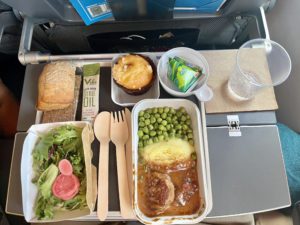 A meal tray on an airplane including peas, vveal, a salad, bread, a muffin, and a water. 