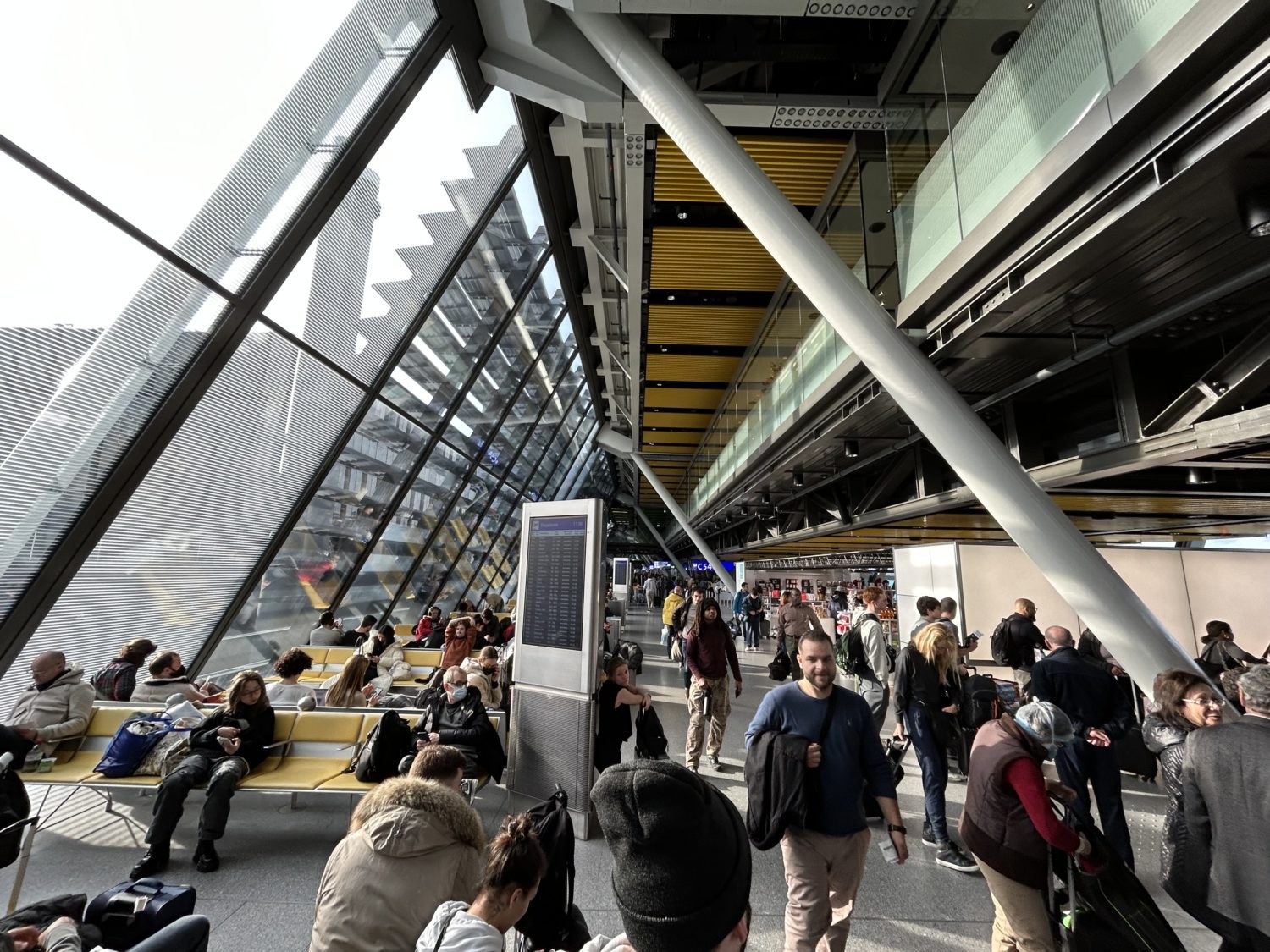 Glassy, sloped ceilings of Geneva Airport as travelers sit and mull around in natural light. 