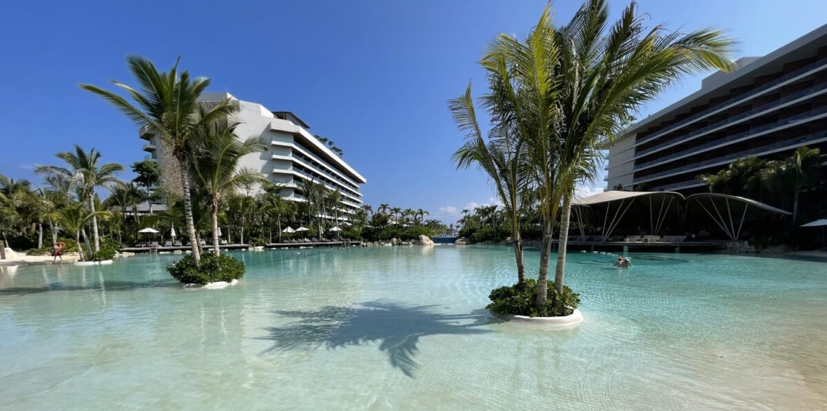 a large, shallow hotel pool with two palm trees on islands in the middle.