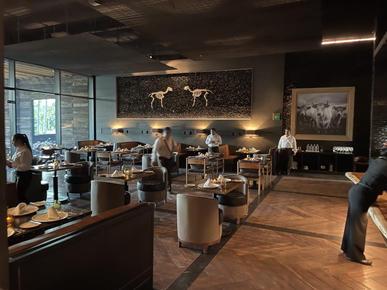 A hotel restaurant with tables for two. There's an art piece on the wall of two animal skeletons. 