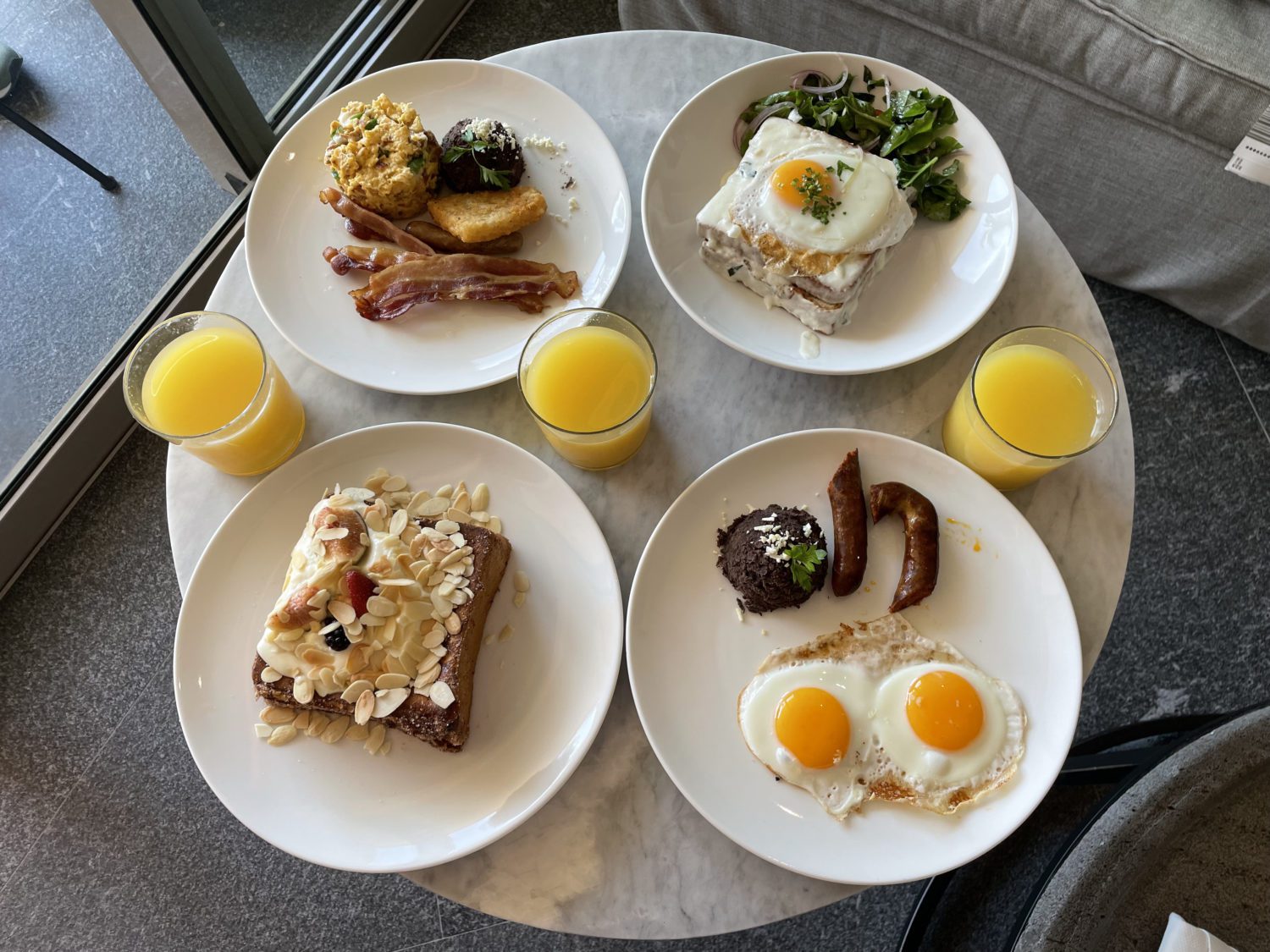 A cafe table with four plates with various breakfast dishes like eggs, french toast, and bacon and three glasses of orange juice. 
