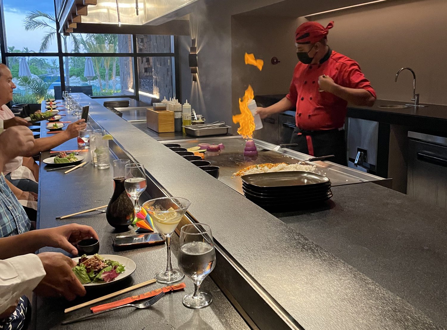 Hibachi at a hotel restaurant. Diners are seated along the counter and the chef is wearing a red chefs coat and bandana as he lights an onion volcano. One customer is filming the scene on their phone. 