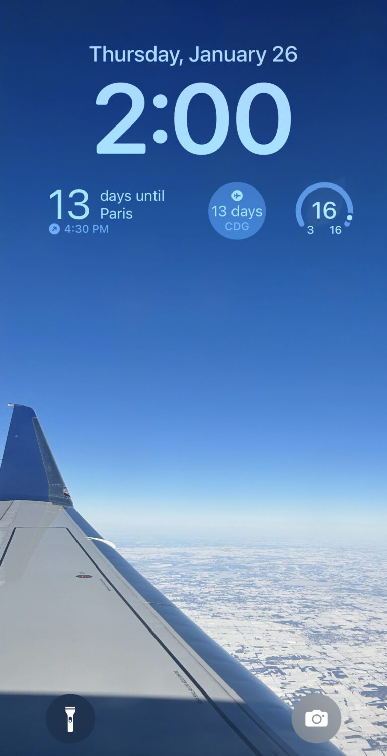 A lock screen with the date, time, and two different Flighty Pro widgets detailing a flight to Paris in 13 days. 