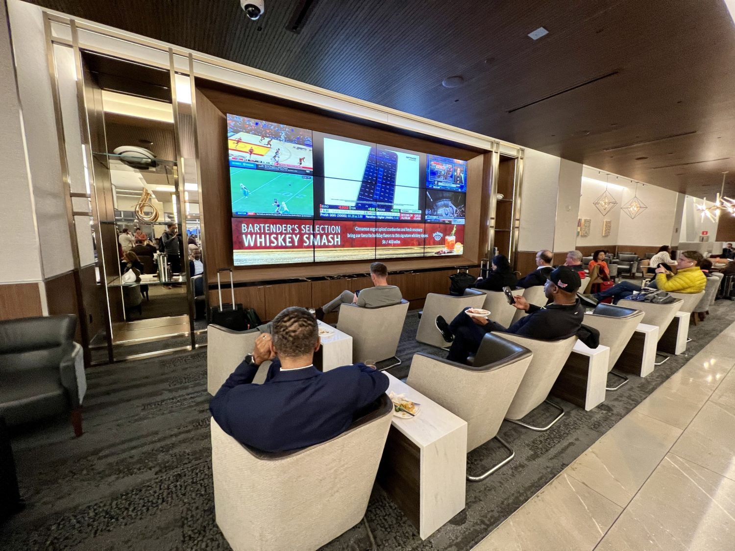 airport lounge guests sitting in small armchairs in front of a large TV