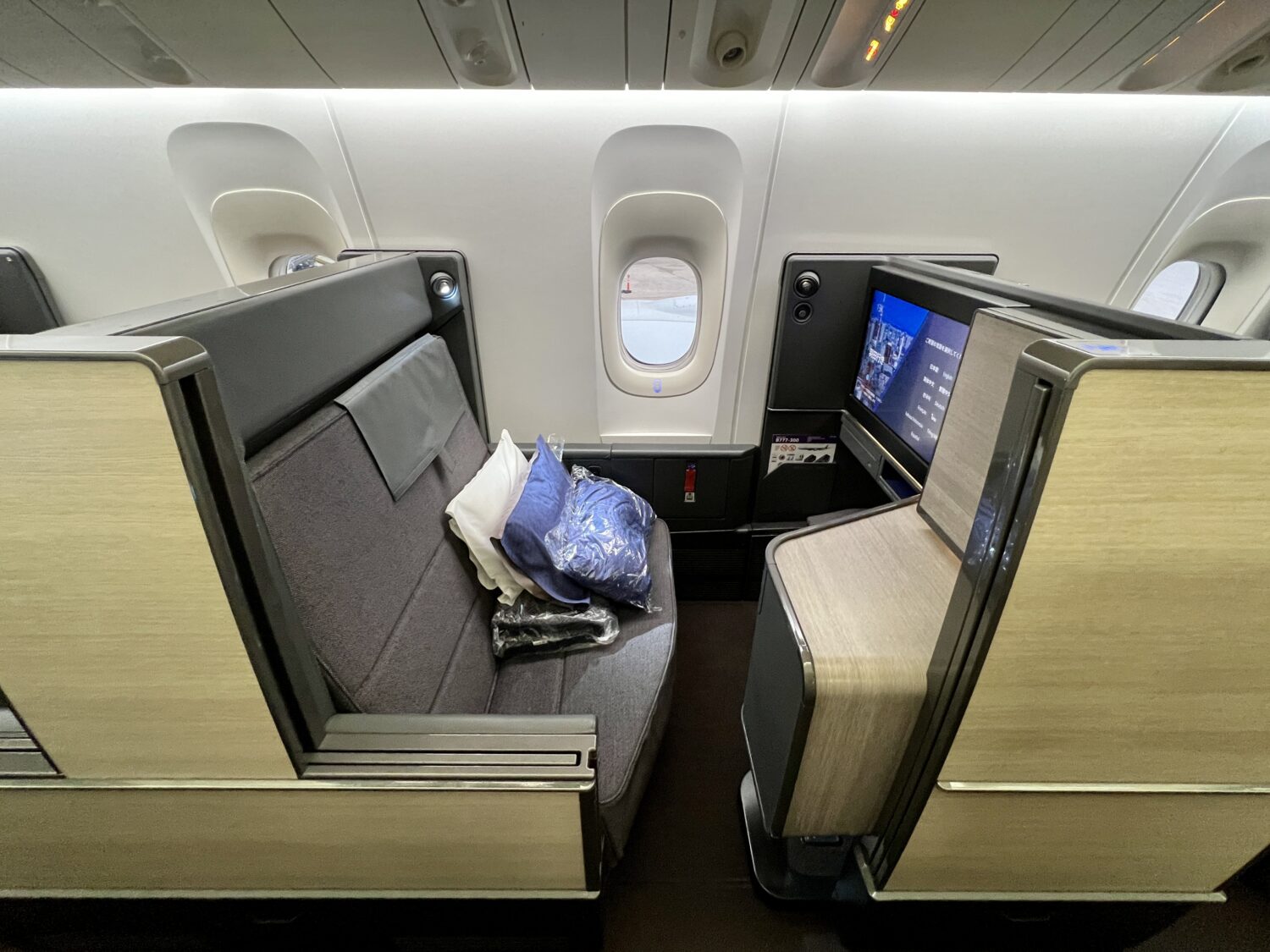 a business class suite with a gray padded seat, light wood walls, and a blanket and pillow on the seat. 