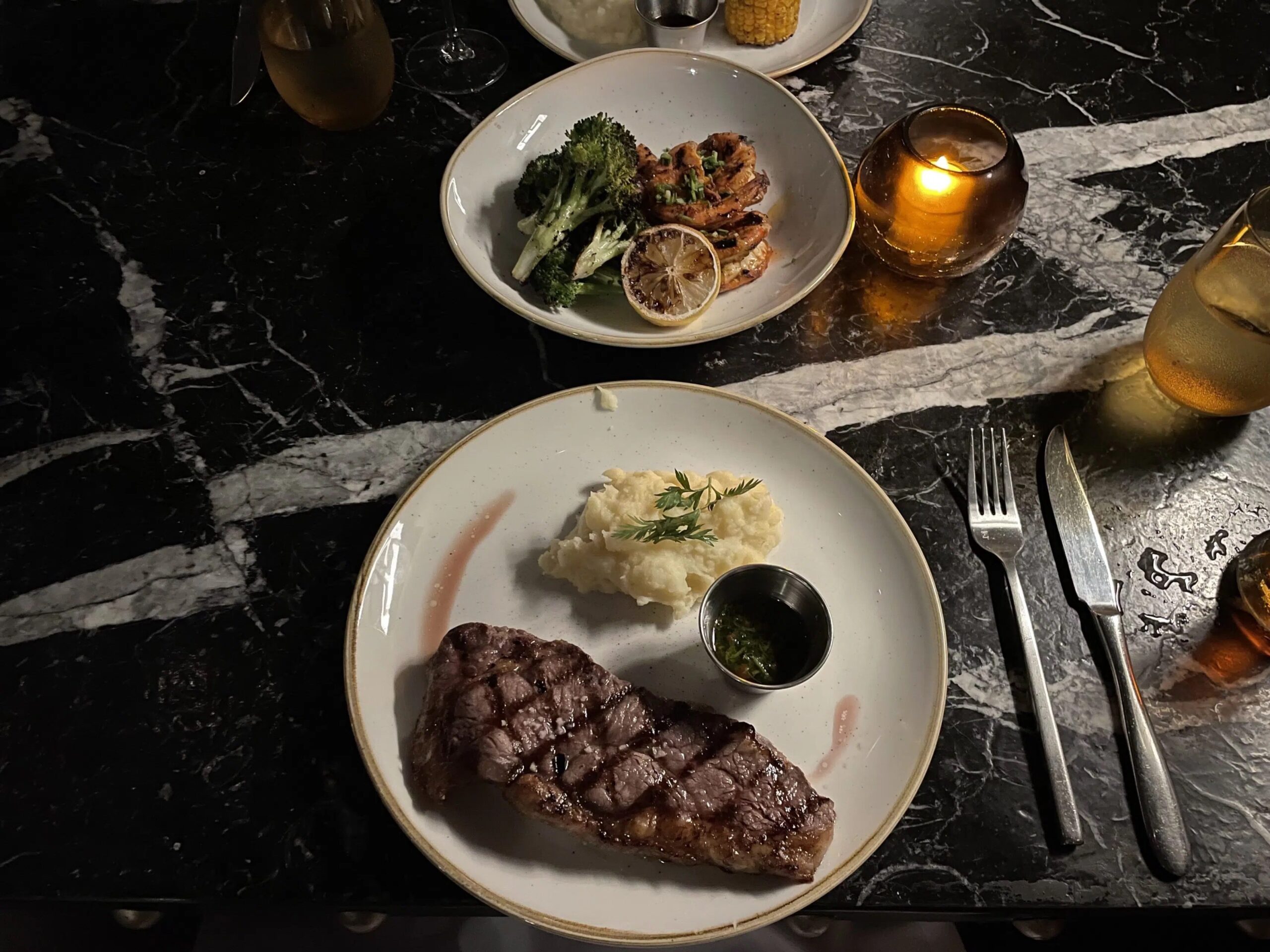 a dinner plate with steak and mashed potatoes and a side plate with broccoli, shrimp, and a lemon slice. 
