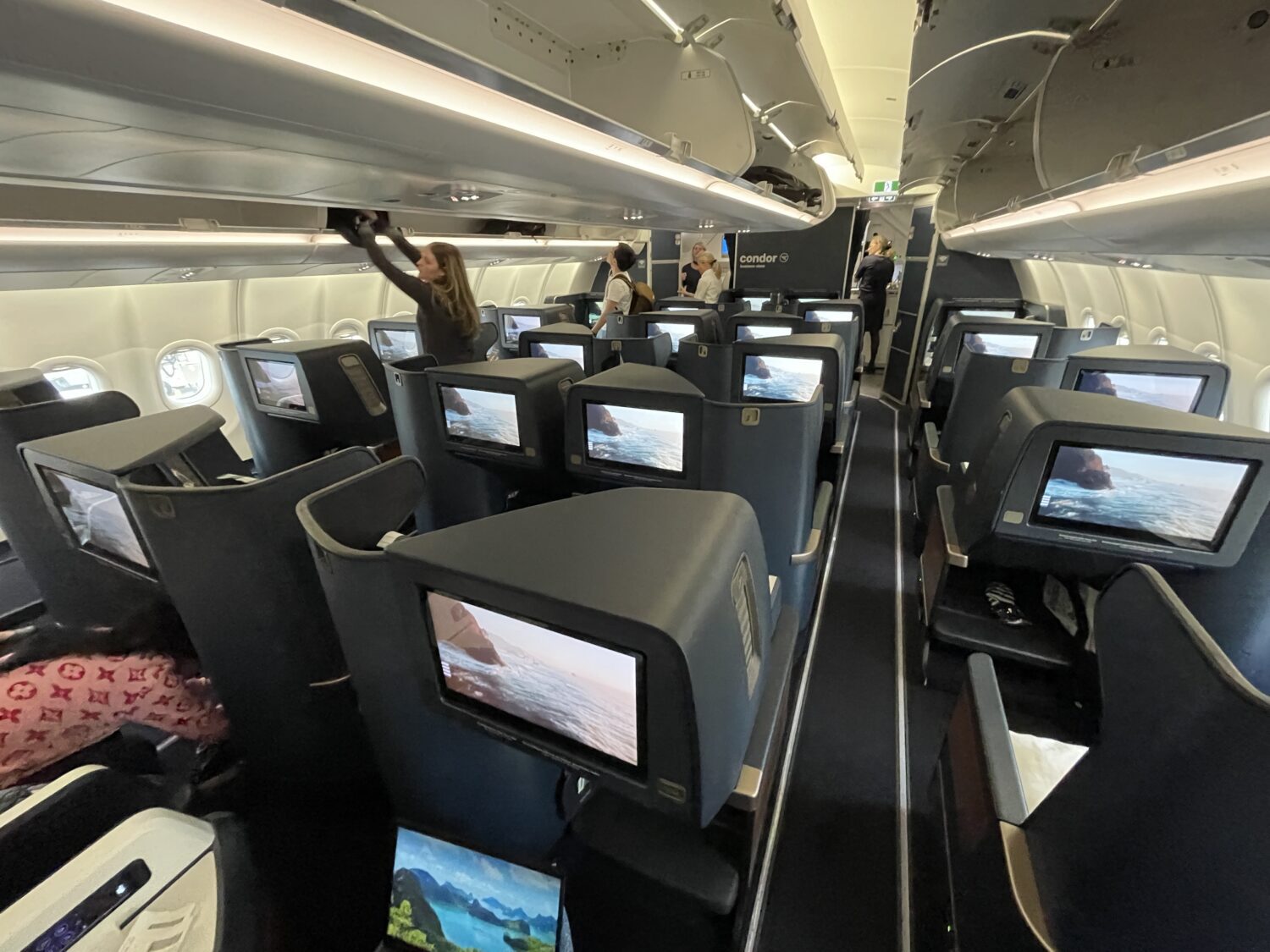rows of business class seats with screens and passengers putting bags in the overhead bins on an airplane