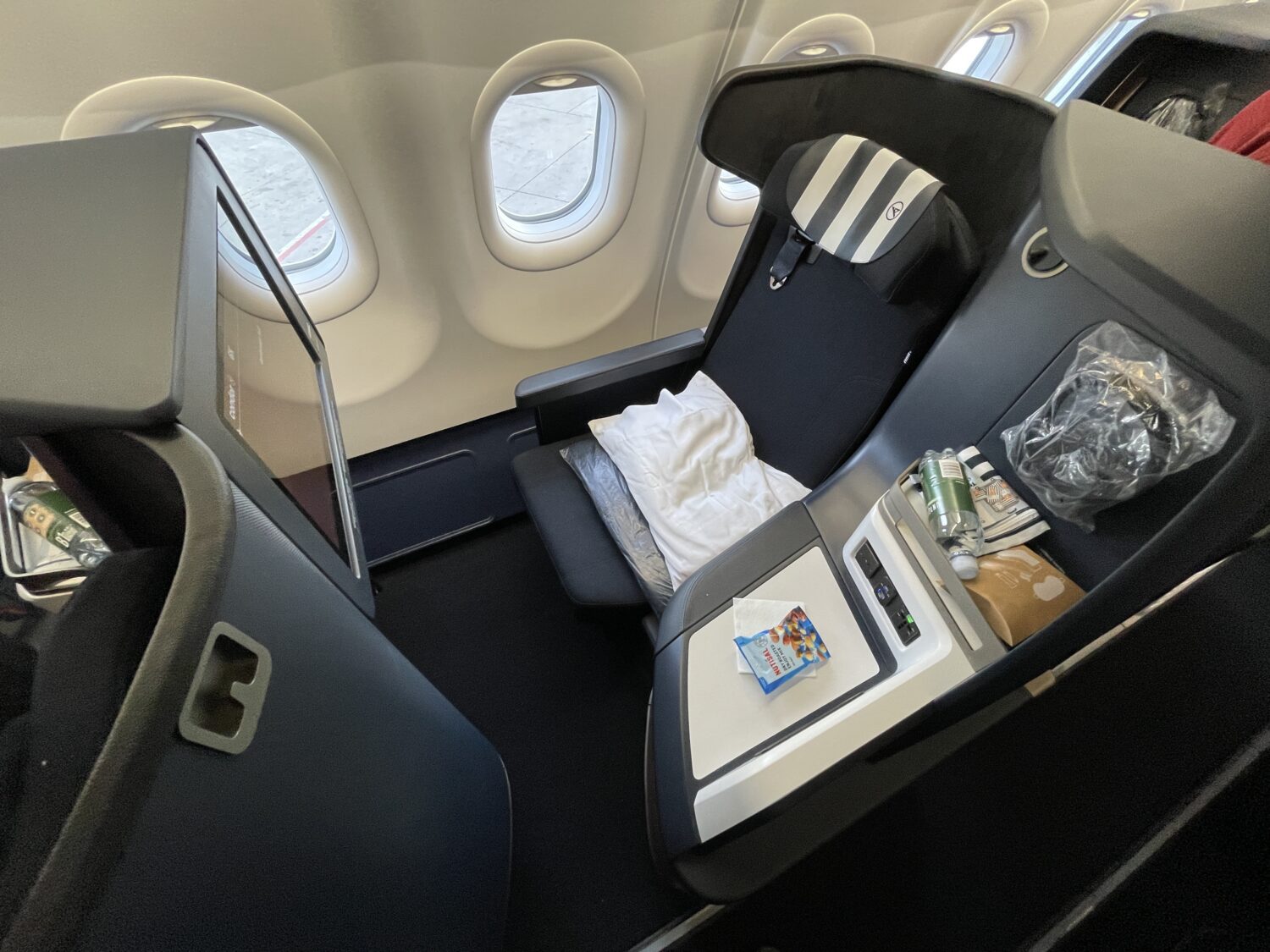 a business class seat with a striped headrest, pillow, snacks, a water bottle, and headphones in a plastic bag
