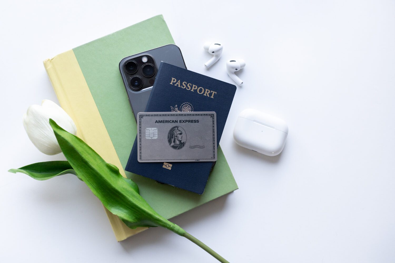 an american express platinum card sitting on top of a book, phone, and passport with a white tulip and set of apple airpods to the side