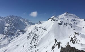 A massive mountain peak, covered in snow, pierces the sky along with a small, single cloud on a sunny day. 