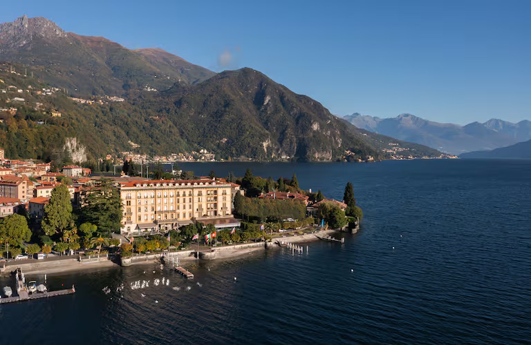 a large hotel building on the shores of a lake with mountains in the background
