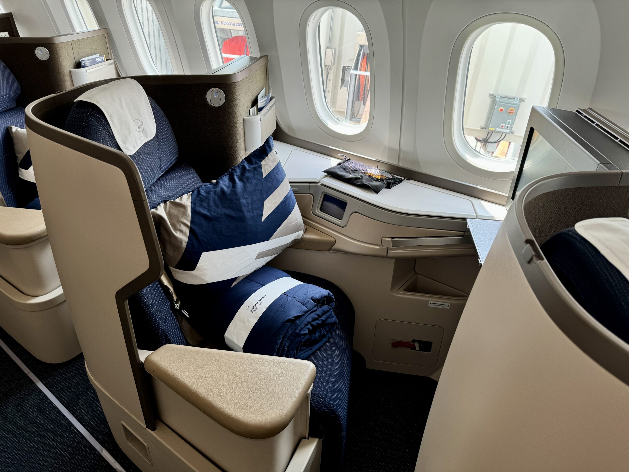 Lufthansa 787-9 business class seat with blue interiors and eggshell shells with a pillow and blanket on the seat. 