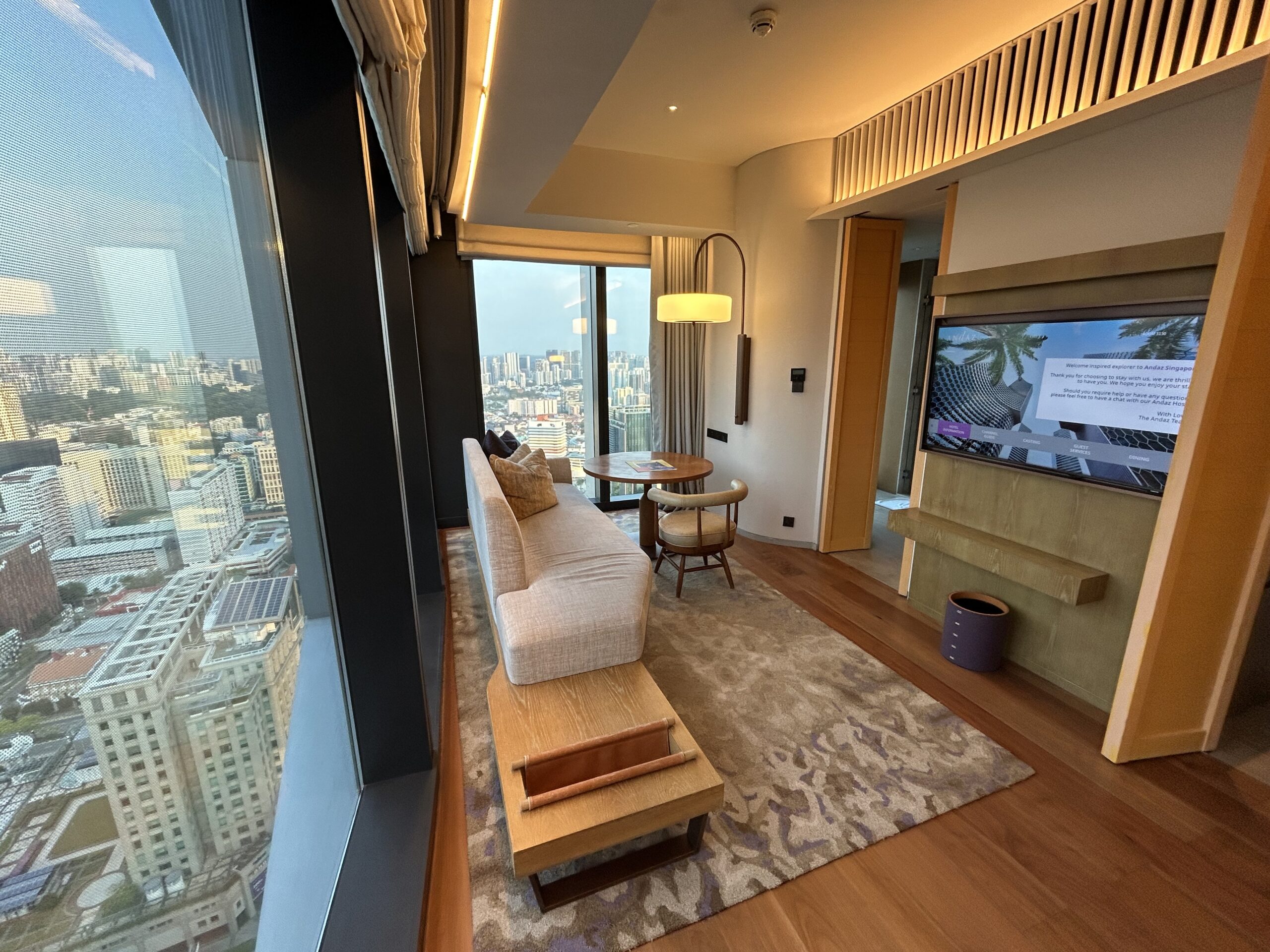 corner room in a hotel with a long couch, TV and city views