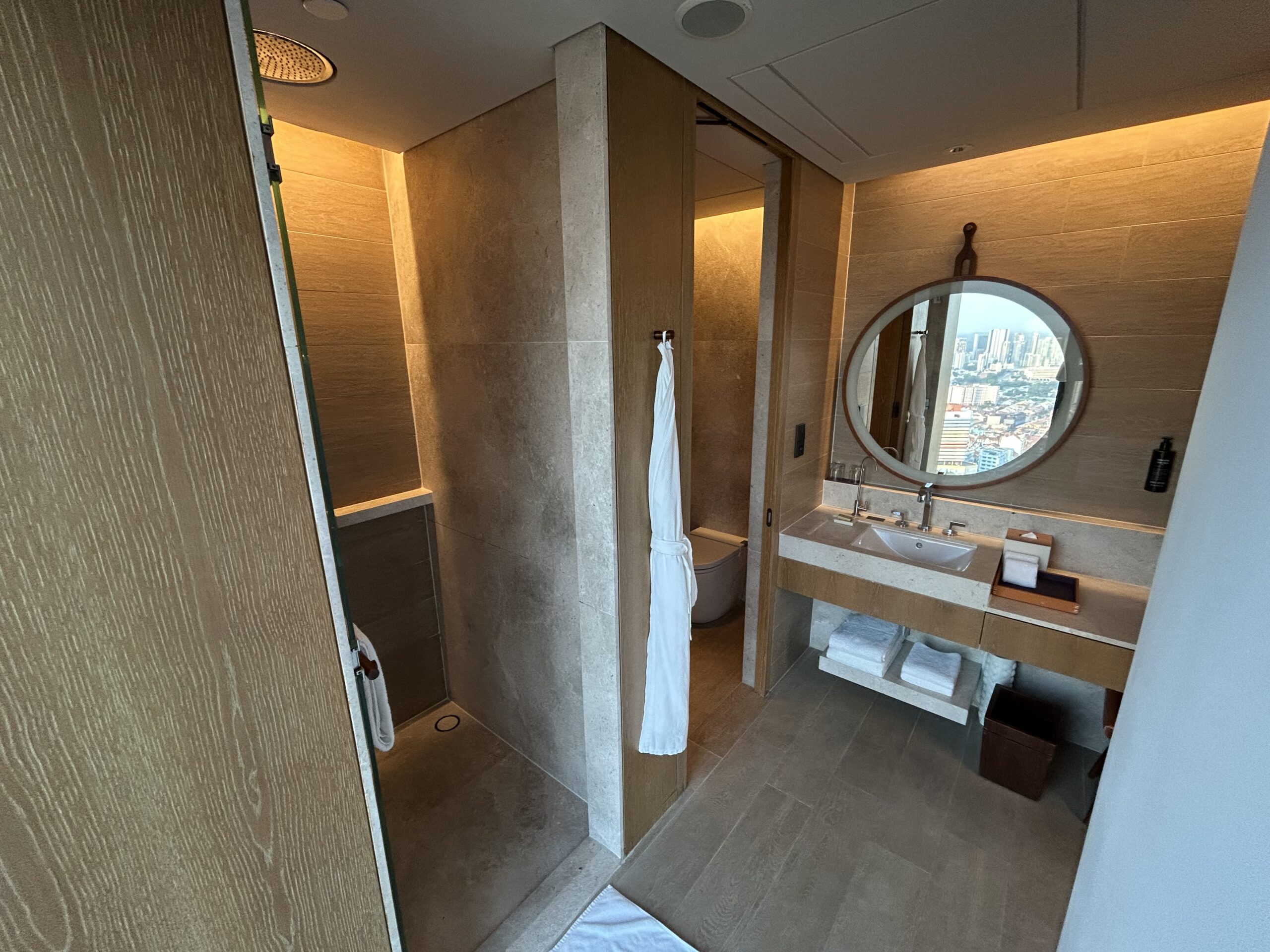 a hotel bathroom with a large circular mirror, sink, and separate room for a toilet. There's a bathroom hanging on a hook on the wall. 