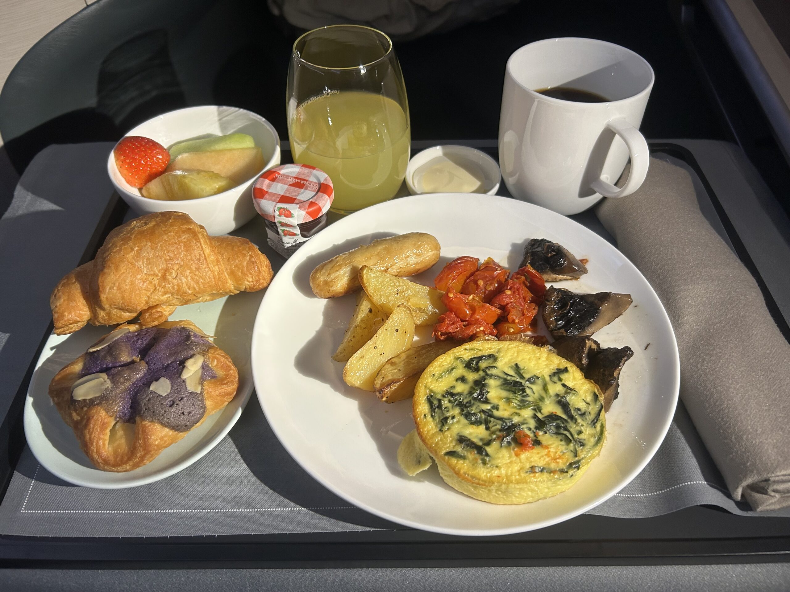 pastries, a bowl of fruit, a glass of juice, cup of coffee, eggs and potatoes served on an airplane tray table. 