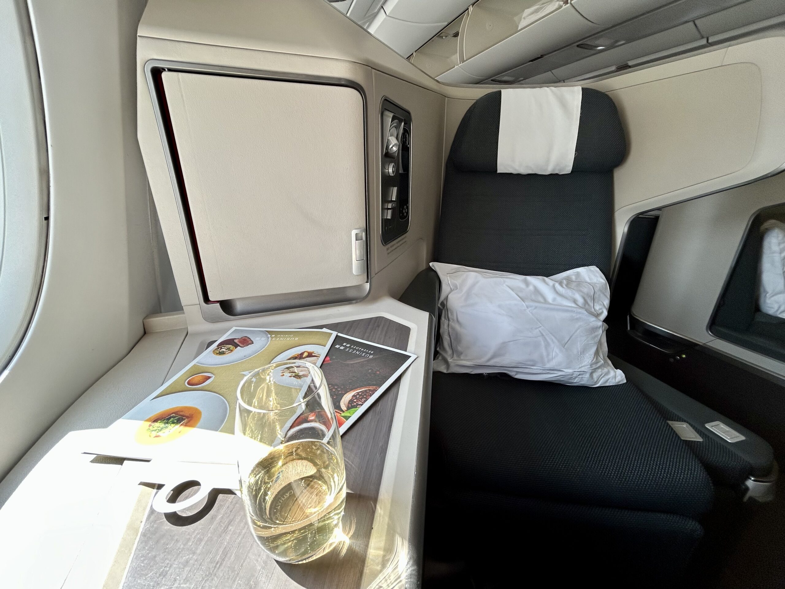 a business class seat with a pillow on the seat. There is a glass of champagne and two menus on the side table. 