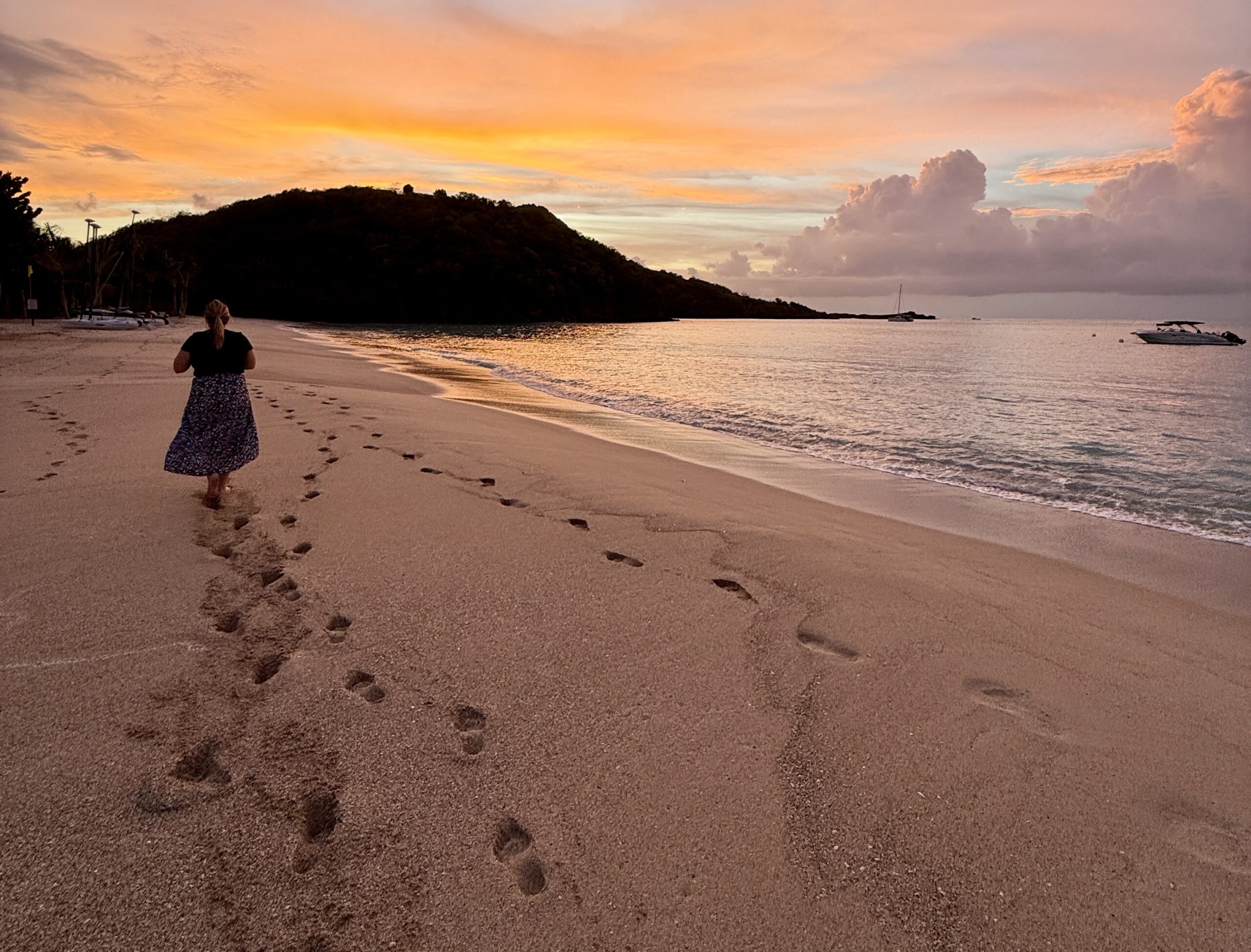 a woman walking down the beach with the sun setting behind a hill
