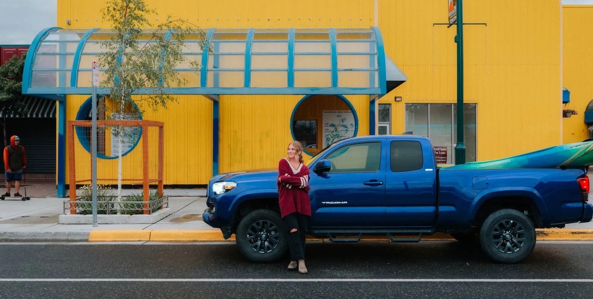 a woman in a red dress leaning against a blue pickup truck in front of a yellow building