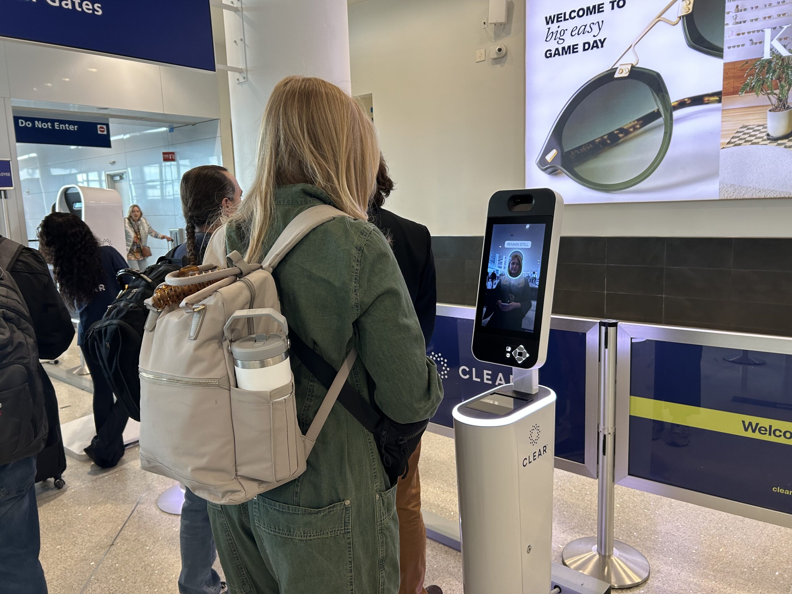 A traveler in a green jumpsuit and a pink backpack gets her face scanned by a CLEAR EnVe pod at the airport. 