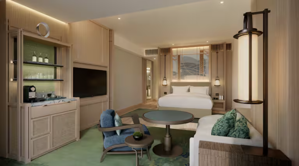 a hotel room with green carpet, a small seating area and a mini bar with an espresso machine. At the back of the room is a queen-size bed with two lamps on either side. 