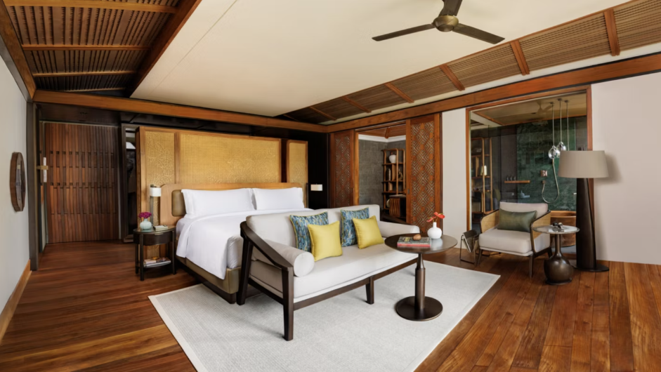 a hotel room with dark wood floors and ceilings, a large bed and small couch with a table. There's an armchair and lamp to the left of the bed. 