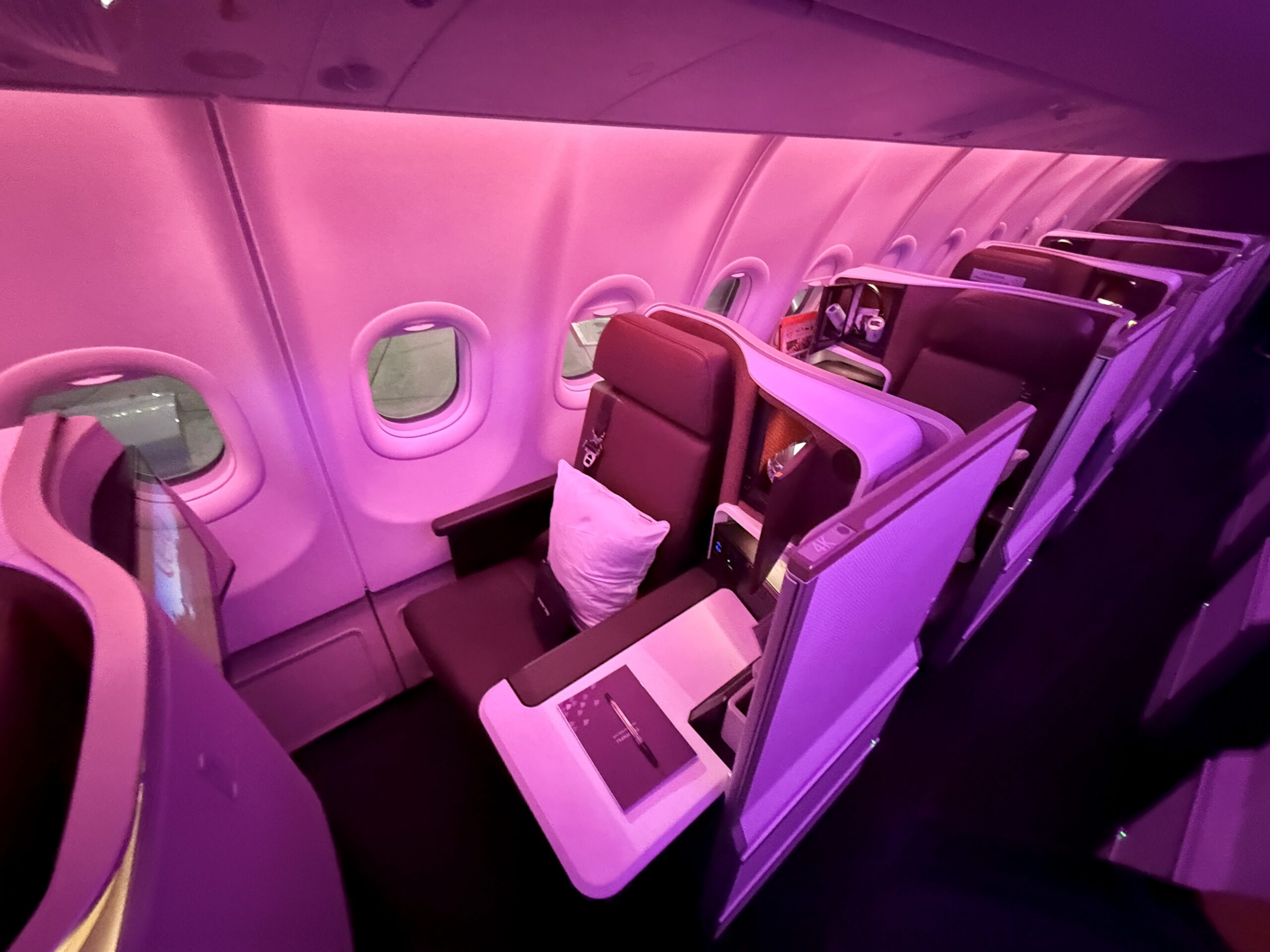 a business class seat on a plane with purple and red lighting. The seat is closer to the window, with a console table on the side closest to the aisle. 